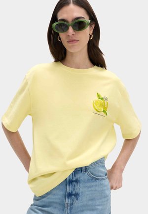 Kvinde iført en lysegul T-shirt med citronmotiv og grønne solbriller, kombineret med blå cowboybukser, poserende med hænderne på hoften.