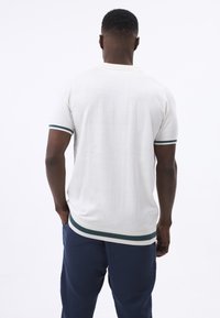 Kortærmet, hvid strikket polo-sweater med mørkegrønne og hvide striber på ærmerne og kanten. Glat tekstur, tætsiddende form.