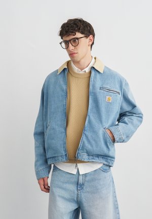 Young man wearing tortoiseshell glasses, beige sweater layered over white shirt, light blue denim jacket and jeans, standing against plain background.