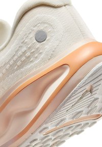 Gros plan sur le talon d'une basket beige avec une tige en tissu texturé, un accent pêche sur la semelle intermédiaire et une semelle extérieure en caoutchouc blanc à motifs.