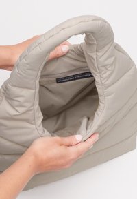 Borsa color beige chiaro con una texture morbida e trapuntata e una maniglia arrotondata, che mostra un ampio interno con un'etichetta che indica il materiale realizzato con reti da pesca riciclate.
