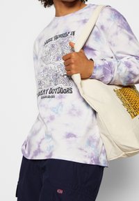 Chemise à manches longues tie-dye en violet et blanc avec un imprimé graphique « Choisis-toi dans la grande nature », associée à un sac fourre-tout beige.