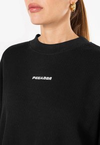 Fekete bordázott pulóver, kerek nyakkivágással és fehér "PEGADOR" logoval. Texturált anyaggal és látható varrási részletekkel rendelkezik.