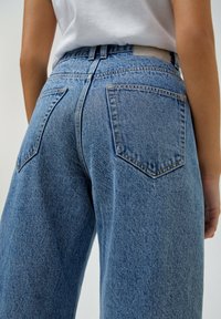 Jeans de denim azul de talle alto con un corte relajado, que presentan costuras visibles, dos bolsillos traseros y un ligero desgaste en la textura de la tela.