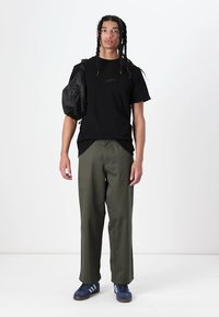 Black t-shirt with stitched lettering, olive green loose-fit pants, and blue sneakers with white stripes. Carrying a black shoulder bag.