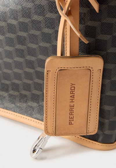 Ετικέτα απο ταμπά δέρμα βαλίτσας με ανάγλυφη την επιγραφή «PIERRE HARDY», προσαρτημένη σε σκούρα τσάντα με ανάγλυφη υφή, επένδυση από ανοιχτό καφέ δέρμα και ασημένιο κούμπωμα.
