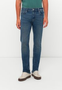 Jeans de mezclilla azul oscuro con corte ajustado, cinco bolsillos y un leve desgaste. Combinados con zapatillas de color claro y una camiseta verde.