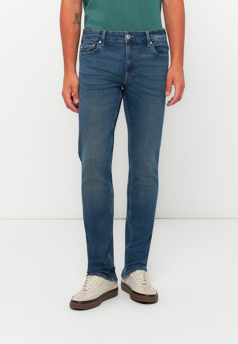 Jeans de mezclilla azul oscuro con corte ajustado, cinco bolsillos y un leve desgaste. Combinados con zapatillas de color claro y una camiseta verde.