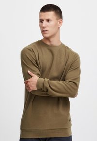 Pull à manches longues vert olive en tissu texturé, avec un col rond, des poignets côtelés et un ourlet droit. Détail logo subtil près de l'ourlet.