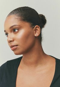 Ørestik med tre klare ædelsten i en lineær rækkefølge, sat i metal med sølvfarve. Modellen viser ørestikkets profil mod en neutral baggrund.