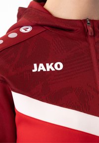 Rode sportjack met een gestructureerde stof, witte accenten en het "JAKO"-logo. Inclusief een ritssluiting op de capuchon en een patroon op de schouder.