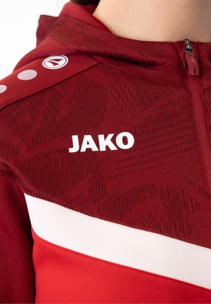 Giacca sportiva rossa con tessuto testurizzato, dettagli bianchi e logo "JAKO". Include un cappuccio con zip e un design a motivi sulla spalla.