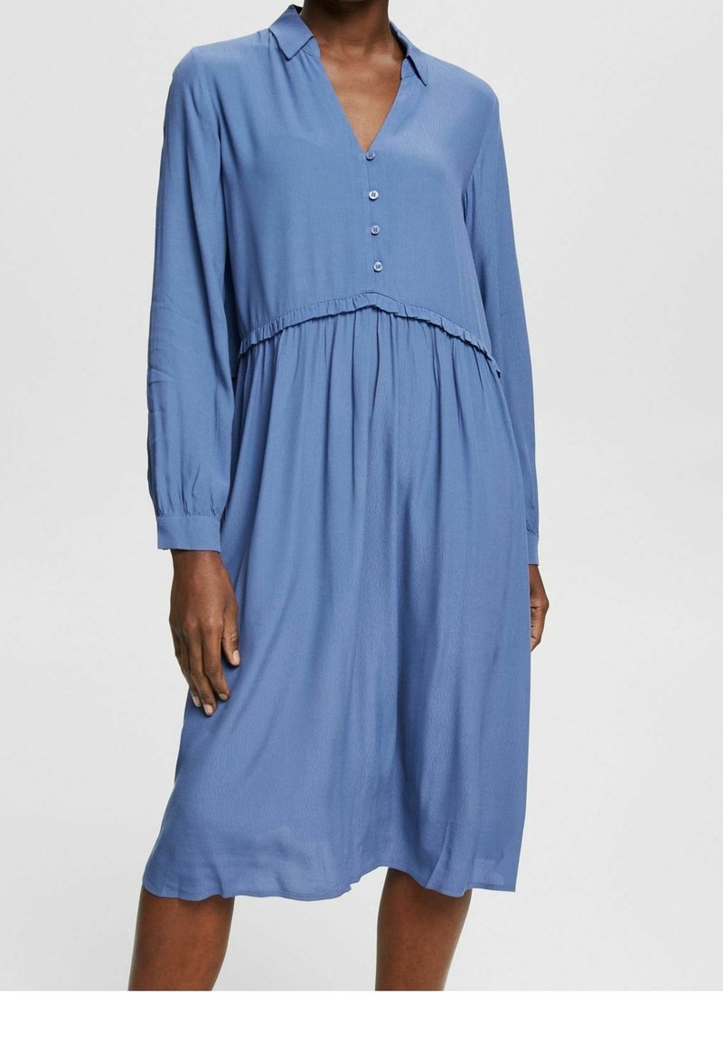 Femme portant une robe midi bleue ample à manches longues, boutonnée, avec une taille froncée et un col en V.