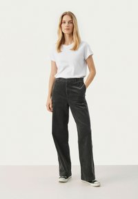 Pantalons en velours côtelé noir à jambe droite, associés à un t-shirt blanc uni. Le mannequin se tient avec les mains dans les poches, portant des baskets noires.
