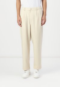 Beige pleated trousers in lightweight fabric with a loose fit and tapered legs, paired with white sneakers; smooth texture and minimal design.