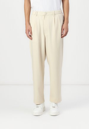 Beige pleated trousers in lightweight fabric with a loose fit and tapered legs, paired with white sneakers; smooth texture and minimal design.