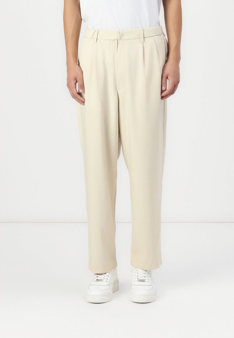 Beige pleated trousers in lightweight fabric with a loose fit and tapered legs, paired with white sneakers; smooth texture and minimal design.