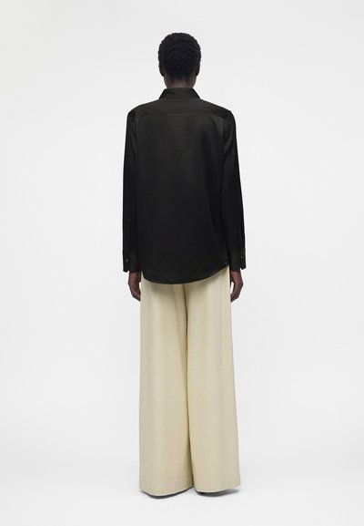 Άτομο που φοράει μαύρο πουκάμισο με μακριά μανίκια και μπεζ παντελόνι με φαρδιά μπατζάκια, όρθιο με την πλάτη προς ένα απλό φωτεινό φόντο.