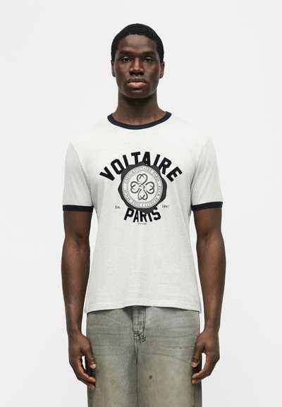 Νέος άνδρας φοράει γκρι ανοιχτό μπλουζάκι Voltaire Paris με μαύρο περίγραμμα και ξεθωριασμένο μπλε τζιν παντελόνι, στέκεται μπροστά σε μια απλή λευκή φόντο.