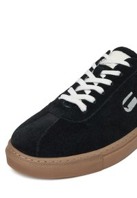 Čierne suede tenisky s bielymi šnúrkami. Majú gumovú podrážku a jednoduché logo. Zaoblená špička s prešitím pozdĺž hornej časti.