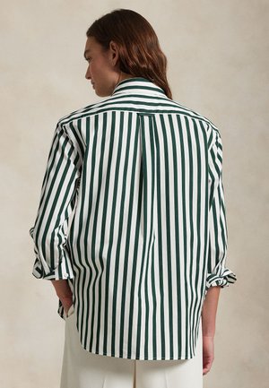 Persoon met lang bruin haar, gekleed in een wit shirt met donkergroene verticale strepen en een witte broek, handen in de zakken, met de rug naar voren.