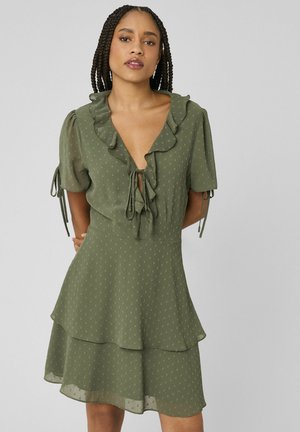 Femme aux cheveux tressés portant une robe vert olive à manches courtes, avec un col à volants et une jupe à couches, debout devant un fond uni.