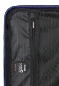 El interior de la funda de viaje azul presenta un bolsillo de malla negra con cremallera, un forro negro suave con líneas sutiles y una etiqueta de marca con instrucciones de cuidado.