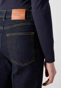 Tmavě modré džíny z denimového materiálu s koženým logem GANT na zadní straně. Mají žluté švy a klasický design s pěti kapsami.