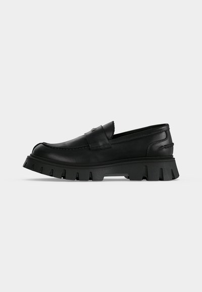 Mocassin en cuir noir avec une semelle épaisse et rainurée ainsi qu’une couture moc toe, vu de profil sur un fond gris clair uni.