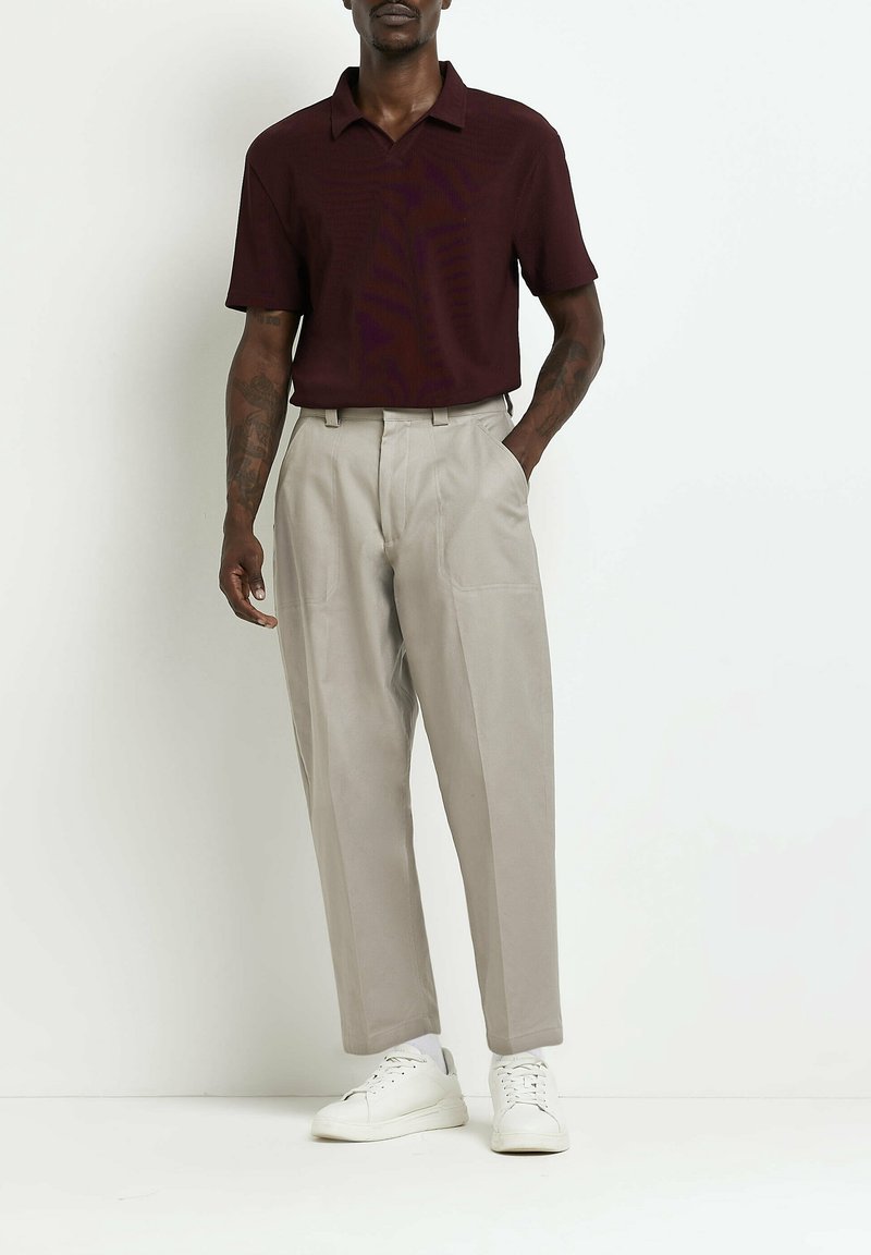 Homme portant un polo à manches courtes foncé, pantalon beige taille haute, baskets blanches, debout avec une main dans la poche sur un fond uni.
