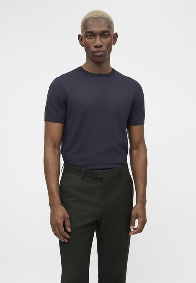 Άνδρας με κοντά ξανθά μαλλιά που φοράει στενή μπλε σκούρα κοντομάνικη μπλούζα και σκούρο πράσινο παντελόνι, στέκεται μπροστά σε απλό φωτεινό φόντο.