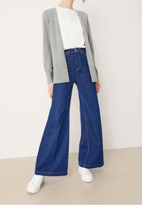 Cárdigan gris de frente abierto, camiseta blanca y jeans de mezclilla oscuros de pierna ancha. Zapatillas con material blanco texturizado. Conjunto simple y casual.