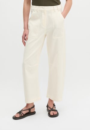 Personne portant un pantalon crème taille haute à jambes larges avec poches plaquées à l'avant, associé à des sandales plates noires à brides et un haut vert foncé.