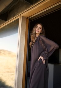 Duga, lepršava haljina od tamnosmeđeg šifona s volanima na rukavima i vezanjem oko vrata, postavljena nasuprot drvenim vratima i pogledom na otvoreno.