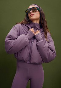 Lila Daunenjacke mit hohem Kragen und Reißverschluss, mit elastischen Bündchen. Getragen mit passenden Leggings. Große, dunkle Sonnenbrille.