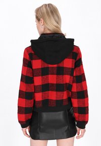 Giacca a quadri rossi e neri tagliata, con cappuccio nero, caratterizzata da una texture morbida e polsini a costine. Coordinata con una gonna di pelle nera.