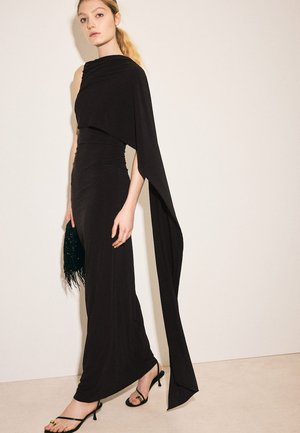 Femeie într-o rochie neagră lungă, cu un umăr gol și mânecă drapată, ținând o geantă neagră cu franjuri, purtând pantofi negri cu barete.