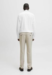 Camicia bianca a maniche lunghe con polsini abbottonati, abbinata a pantaloni beige con due tasche posteriori, vista da dietro. Mostrati scarpe nere.