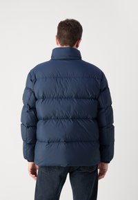 Mörkblå puffjacka med hög krage, med horisontella quiltade sektioner och elastiska manschetter. Buren över mörka jeans.