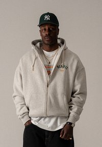 Sivá mikina na zips s predným vreckom, na ktorej je vyšitý nápis "BLIVIS NY" v zelenej a oranžovej farbe. Nosená cez biele tričko s potlačou.