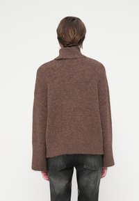 Pull en tricot marron avec un col haut, offrant une coupe décontractée, des manches longues et un ourlet légèrement effiloché, associé à un jean foncé.