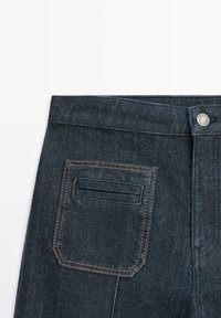 Dunkelblaue Jeans aus Denim mit Knopfverschluss, Gürtelschlaufen und einer einzelnen Gesäßtasche mit orangefarbener Naht. Glatte Textur.