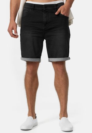 Homme portant un short en denim noir retroussé, main dans la poche, et des chaussures blanches sans lacets, cadré à la taille et sous les genoux.