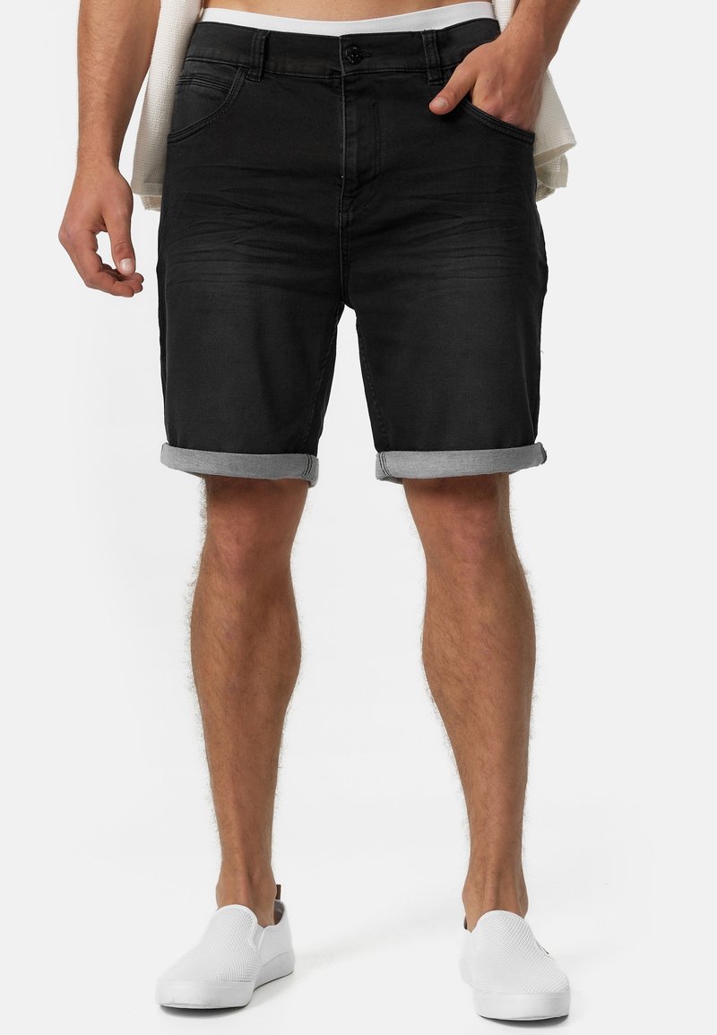 Homme portant un short en denim noir retroussé, main dans la poche, et des chaussures blanches sans lacets, cadré à la taille et sous les genoux.