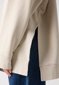 Beige langwerpig shirt met ribbelstructuur, zijsplitsingen en contrasterende stiksels, gecombineerd met donkere denim jeans.