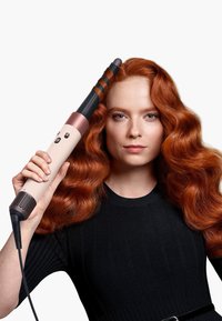 Locktång med svart fat, rosa mittendel och texturerat vitt handtag. Modellen har vågigt röd hår och bär en svart ribbad tröja.