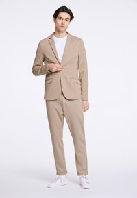 Beżowa kurtka z dwoma przednimi kieszeniami, w zestawieniu z dopasowanymi spodniami. Model nosi białą koszulkę i białe sneakersy.