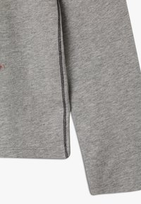 Šedá mikina z materiálu s měkkou texturou, s černým šitím podél švů a malým designovým detailem na přední straně.