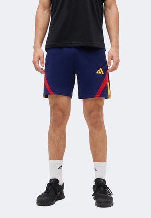 Férfi, aki tengerészkék sportos sortot visel piros és sárga részletekkel, fekete tornacipővel, fehér zoknival és fekete pólóban áll egyenesen.