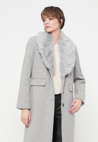 Abrigo de lana gris con cuello de piel sintética, con apertura frontal, dos bolsillos delanteros y una silueta relajada. El modelo lleva pantalones oscuros debajo.